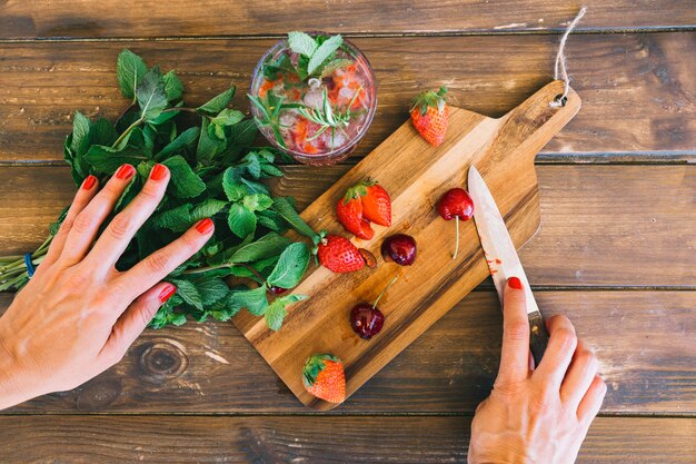 Vista elevada de la mano de la mujer con hoja de menta; jugo; fresas y cerezas en el escritorio de madera