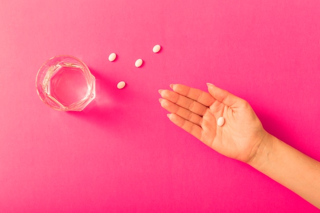 Vista elevada de la mano de la mujer tomando medicina con vaso de agua sobre fondo rosa