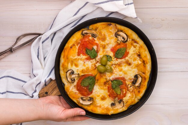 Una vista elevada de la mano que sostiene la pizza hecha en casa sobre el escritorio de madera