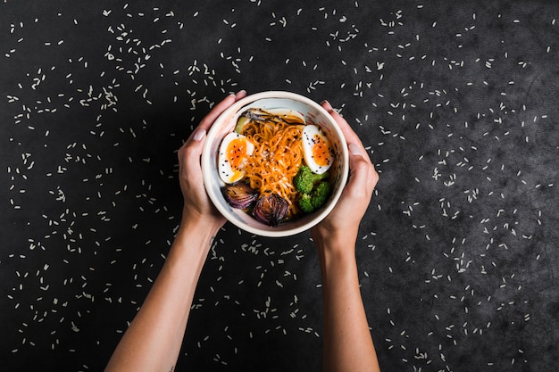 Una vista elevada de las manos que sostienen tazones de fideos ramen con huevos y ensalada con granos de arroz sobre fondo negro