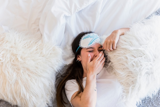 Una vista elevada de la mujer con máscara de ojo acostado en la cama bostezando