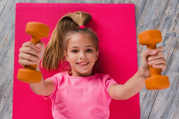 Una vista elevada de una niña rubia acostada en la alfombra rosa haciendo ejercicio con mancuernas