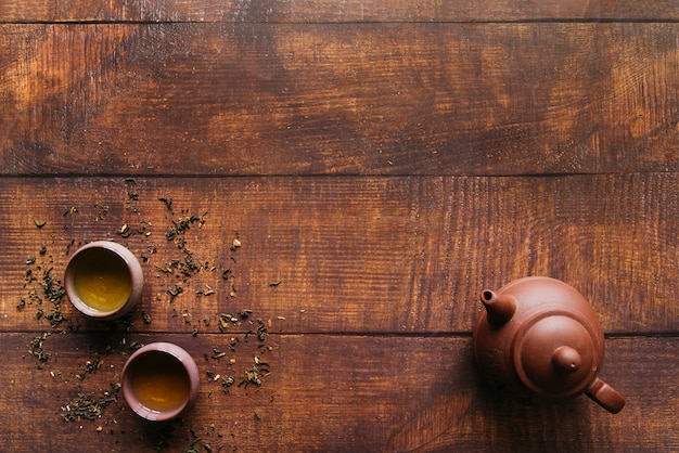 Una vista elevada de la olla de barro con tazas de té de hierbas en el escritorio de madera