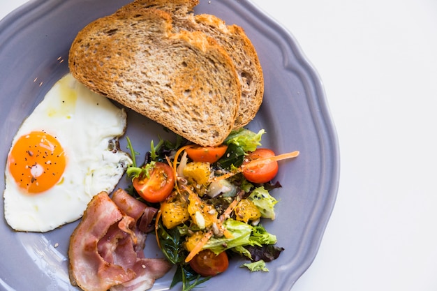 Una vista elevada de pan tostado; huevo frito; tocino; Ensalada en placa gris sobre fondo blanco