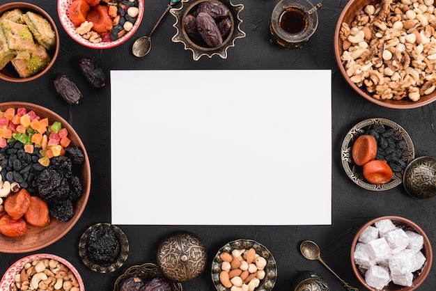 Foto gratuita una vista elevada de papel blanco en blanco rodeado de deliciosas frutas secas; tuercas y dulces para ramadán sobre fondo negro con textura