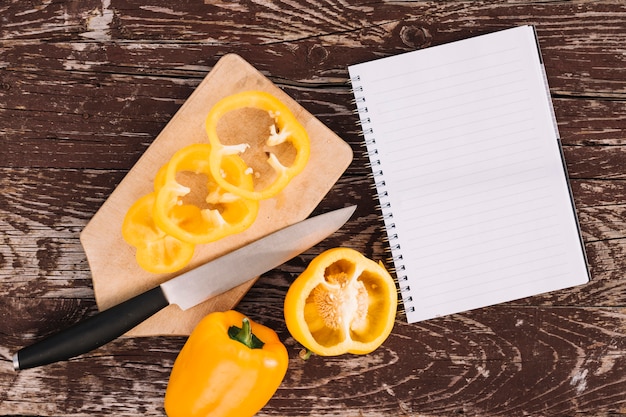 Foto gratuita una vista elevada de pimiento amarillo en tabla de cortar con cuchillo y cuaderno espiral en escritorio de madera