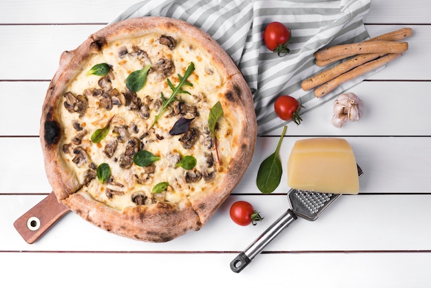 Vista elevada de pizza de champiñones caseros y palitos de pan con ingredientes en un tablón blanco