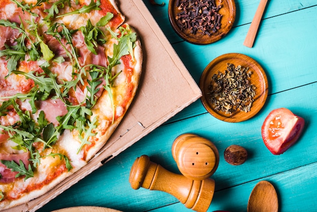 Vista elevada de la pizza italiana del tocino con las hierbas; rodaja de tomate Dientes y peppermill sobre fondo turquesa.