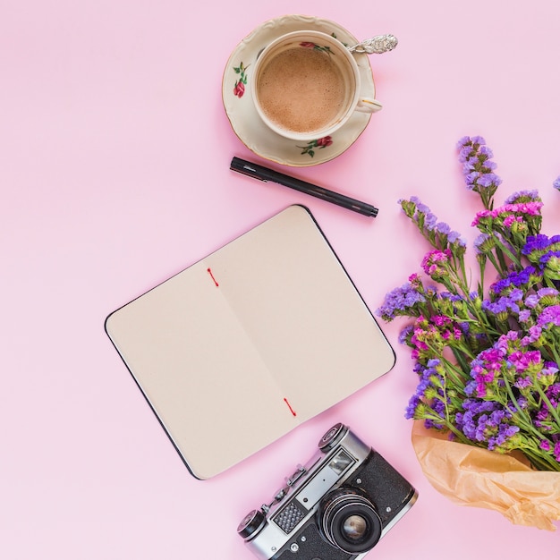 Una vista elevada del ramo de flores; camara vintage diario; Taza de café y pluma sobre fondo rosa