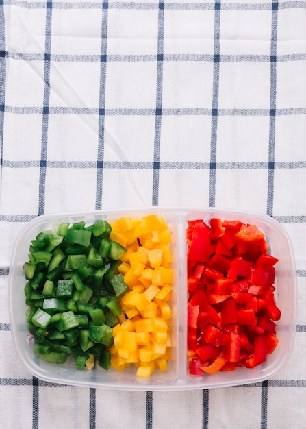 Foto gratuita una vista elevada de rojo picado; pimiento verde y amarillo en el recipiente de plástico sobre el mantel a cuadros