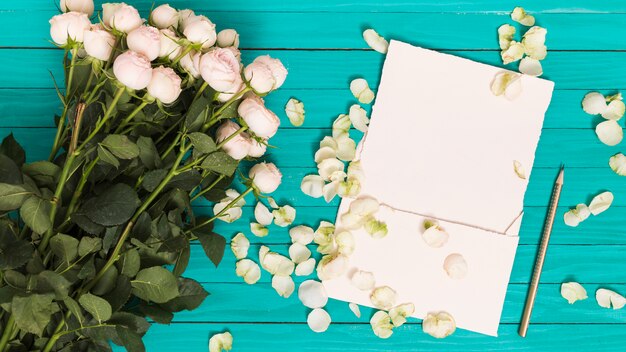 Vista elevada de rosas blancas; lápiz; Hoja en blanco; y pétalos contra mesa de madera verde