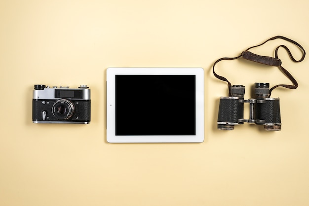 Una vista elevada de la tableta digital con cámara retro y binoculares sobre fondo beige
