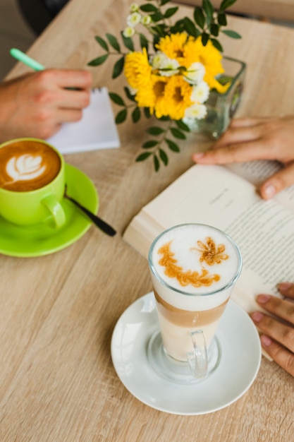 Vista elevada de la taza de café y el café con leche cerca de las personas que estudian en cafés