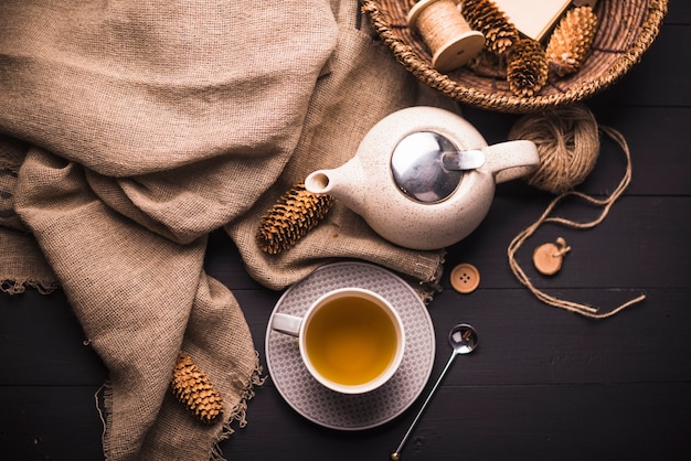 Vista elevada de té de hierbas; piña; tetera; saco; botón; Cesta de mimbre y ovillo de hilo en mesa.