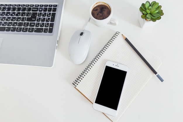 Vista elevada del teléfono celular en el cuaderno y lápiz con la computadora portátil sobre el escritorio blanco