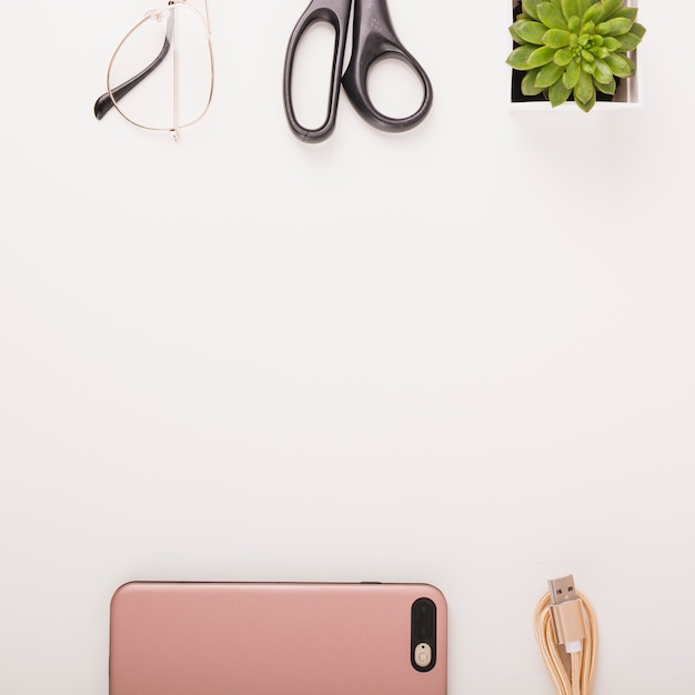 Vista elevada del teléfono inteligente; cable USB; planta en maceta; tijeras y gafas sobre fondo blanco