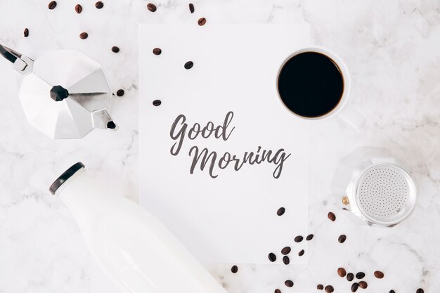 Una vista elevada del texto de buenos días sobre papel; Cafetera Cafetera; taza de café; botella de leche y granos de café sobre fondo de mármol