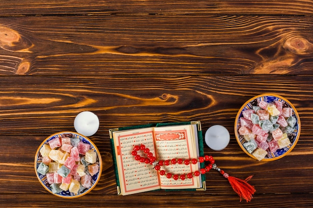 Una vista elevada de las velas; Granos de oración rojos con los tazones de fuente sagrados kuran y lukum en mesa de madera