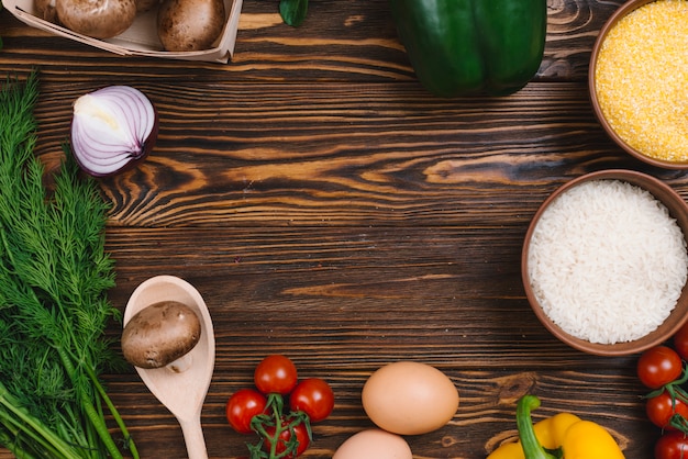 Una vista elevada de verduras con un tazón de granos de arroz y polenta en mesa de madera