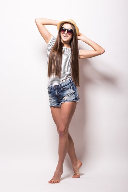 Vista de estudio de mujer alegre con sonrisa dentuda, lleva gafas de sol de moda, sombrero de paja, vestido de verano, aislado sobre una pared blanca