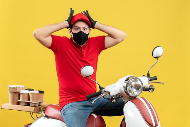 Vista frontal del adulto joven sorprendido vistiendo blusa roja y guantes de sombrero en máscara médica entregando orden sentado en scooter sobre fondo amarillo