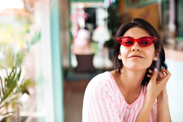 Vista frontal de la agradable mujer bronceada con gafas de sol. Tiro al aire libre de hermosa mujer morena sobre fondo borroso.