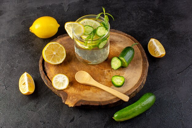 Una vista frontal de agua con limón bebida fresca fresca dentro de un vaso con hojas verdes con cubitos de hielo con rodajas de limones pepino en la oscuridad