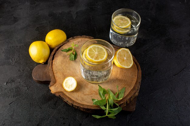 Una vista frontal de agua con limón bebida fresca fresca con limones en rodajas junto con limones enteros y hojas dentro de vasos transparentes en la oscuridad