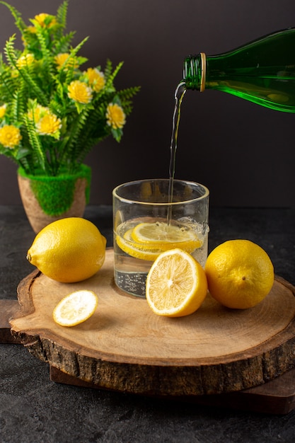 Una vista frontal de agua con limón fresca bebida fría que vierte en el vaso con limones en rodajas junto con limones enteros y flores en la oscuridad