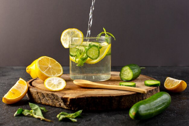 Una vista frontal de agua con limón refresco fresco que vierte dentro del vaso con hojas verdes con cubitos de hielo con rodajas de limones pepino en la oscuridad