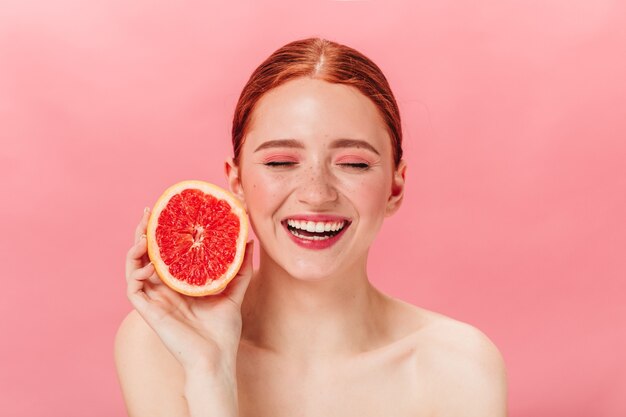 Vista frontal de la alegre chica desnuda con pomelo fresco. Foto de estudio de mujer de jengibre sonriente entusiasta con cítricos.