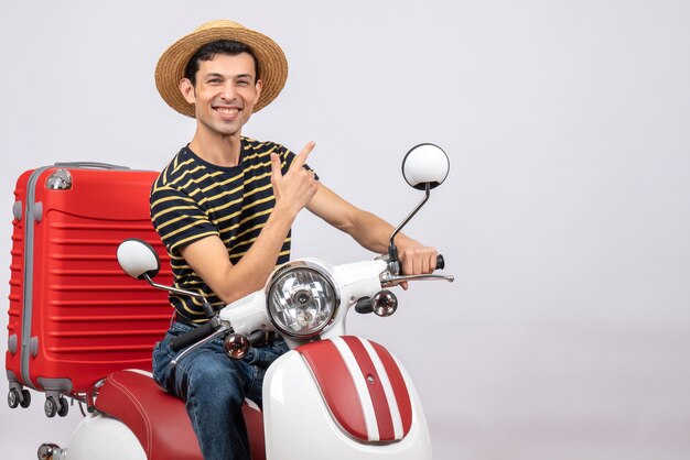 Vista frontal del alegre joven con sombrero de paja en ciclomotor apuntando a algo