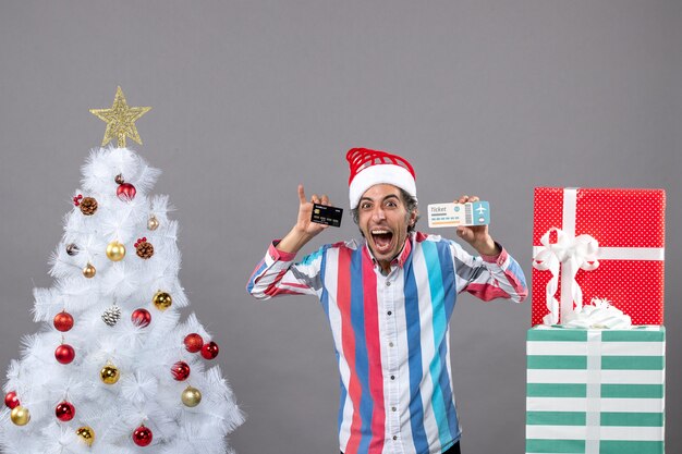 Vista frontal alegre joven sosteniendo la tarjeta y el billete de viaje alrededor del árbol de Navidad y regalos
