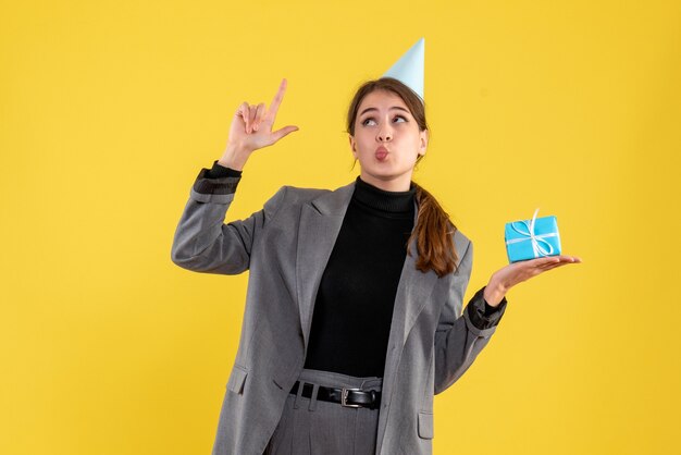 Vista frontal alegre niña con gorro de fiesta sosteniendo su regalo apuntando con el dedo hacia arriba