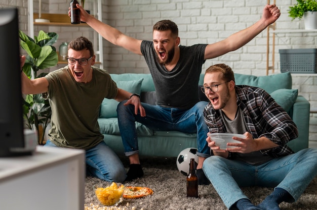 Vista frontal de alegres amigos varones viendo deportes en la televisión juntos mientras toman bocadillos y cerveza