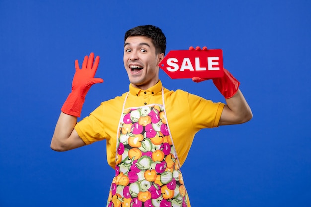 Vista frontal del ama de llaves masculino lleno de alegría con guantes de drenaje con cartel de venta rojo en la pared azul