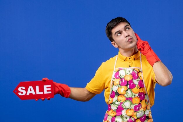 Vista frontal del ama de llaves masculino pensativo en camiseta amarilla con cartel de venta en la pared azul