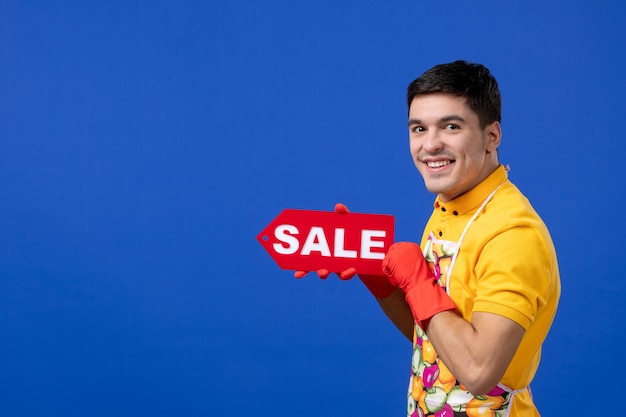 Vista frontal del ama de llaves masculino sonriente en camiseta amarilla con cartel de venta en pared azul aislada