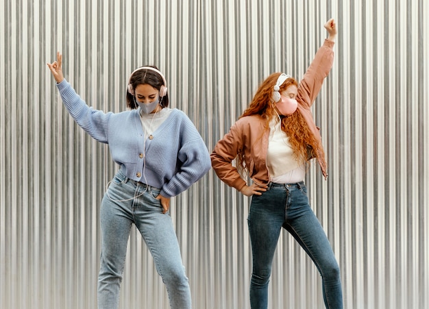 Vista frontal de amigas con máscaras faciales al aire libre bailando con auriculares