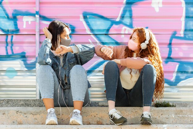 Vista frontal de amigas con máscaras faciales al aire libre escuchando música con auriculares y haciendo el saludo del codo