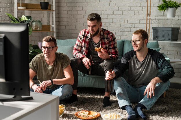 Vista frontal de amigos varones comiendo pizza con cerveza y viendo deportes en la televisión