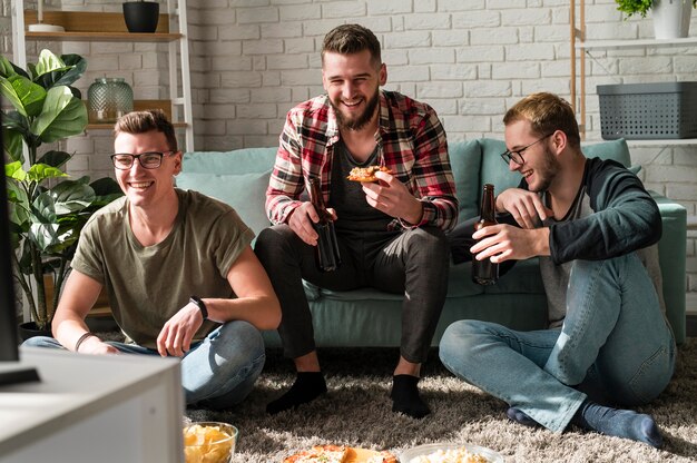 Vista frontal de amigos varones sonrientes comiendo pizza y viendo deportes en la televisión con cerveza