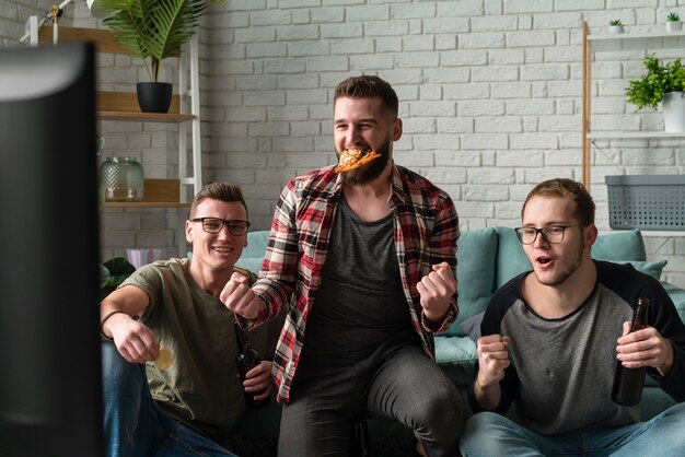 Vista frontal de amigos varones viendo deportes en la televisión y comiendo pizza