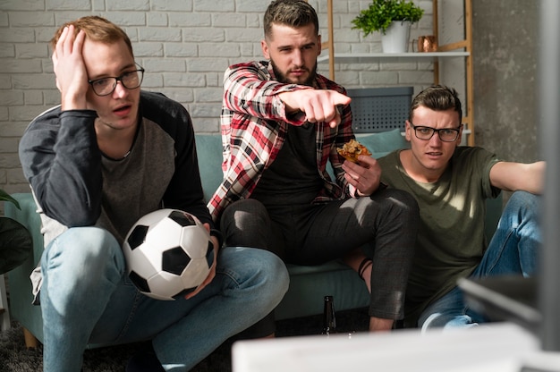 Vista frontal de amigos varones viendo deportes en la televisión con pizza y fútbol