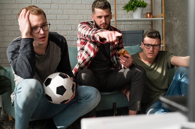 Vista frontal de amigos varones viendo deportes en la televisión con pizza y fútbol