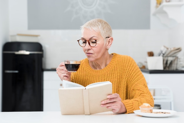 Vista frontal de una anciana leyendo un libro y sosteniendo la taza de café