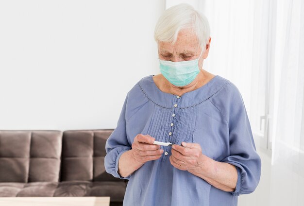 Vista frontal de la anciana con máscara médica mirando el termómetro