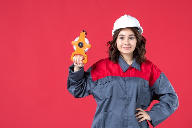 Vista frontal de la arquitecta confía en uniforme con casco sosteniendo cinta métrica en la pared roja aislada