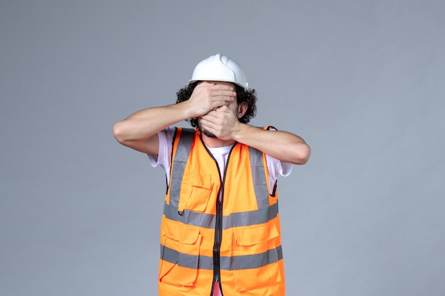 Foto gratuita vista frontal del arquitecto masculino en chaleco de advertencia con casco de seguridad cerrando su rostro en la pared de onda gris