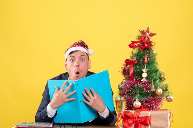 Vista frontal asombrado hombre de negocios sentado en la mesa cerca del árbol de Navidad y presenta sobre fondo amarillo